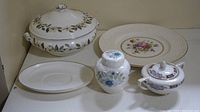 Wide view of five Wedgwood porcelain pieces arranged on a white surface: a large lidded tureen, a floral decorated plate, an oval platter, small floral jar with lid, and sugar bowl with lid