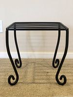 Side view of black metal stool showing curled legs and slatted top surface against beige carpet and wall.