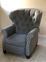 Front view of gray tufted wingback recliner showing upholstery, tufting, nailhead trim, rolled arms, bun feet