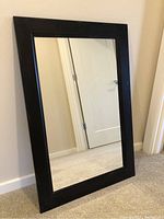 Full view of rectangular mirror leaning against a wall, showing the full black frame and mirror glass.