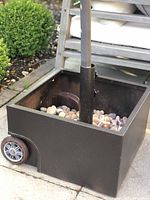 Side view showing brown metal patio umbrella stand with 2 wheels on one side and central pole holder, containing landscaping stones.