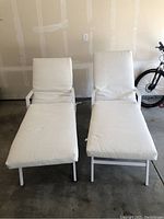Two white metal frame patio loungers with cream cushions placed upright showing shape and size, with bicycle in background.