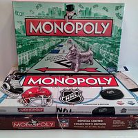 Photo showing three Monopoly board games: Regular Monopoly sealed with green-themed box, NHL Monopoly Collector's Edition with hockey-themed white box, and NFL Monopoly Official Limited Collector's Edition with burgundy-themed box.