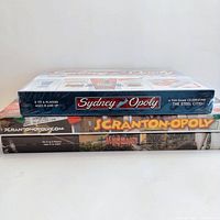 Side view showing the three sealed Monopoly board games stacked, noting titles Sydney Opoly, Scranton Monopoly, and Mississauga Monopoly.