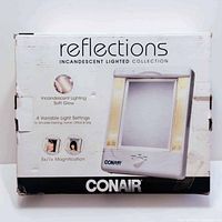 Front view of the Conair Reflections lighted mirror box showing product branding and key features like incandescent soft glow, 4 variable light settings, and magnification.