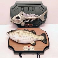 Both novelty singing plaques shown: Big Mouth Billy Bass mounted on wood plaque and Big Mouth Billy Bones skeleton fish on R.I.P. tombstone plaque. Mouths move when singing, bodies and tails do not.