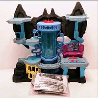Front view of the Batman Batcave playset showing rocky cave design with platforms, central cylinder chamber with Bat emblem, yellow cage, and sealed figure package in front.