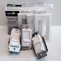 Three Leviton Decora SureSlide dimmer switches shown with packaging and switch plates. Two dimmers still in packaging, one used with missing screw.