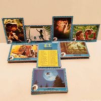 Photo of eight vintage E.T. trading cards arranged on a surface. Cards show colorful images from the E.T. movie including character portraits and iconic scenes. One card is a checklist card showing the complete list of cards from the set.