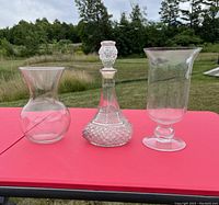 Three glass items displayed: pink flared glass vase, cut-glass style clear decanter with stopper, and a clear pedestal vase.