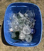 Blue plastic bin holding an assortment of clear glassware items including various bowls, vases, drinking glasses, a small blue and white ceramic dish, and decorative glass plates. Items appear used but in good condition with no visible major damage.