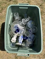 Green storage tub containing an assortment of used glassware and ceramic pieces including candlesticks, bowls, bottles, and glasses.