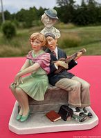 View of the full porcelain figurine showing three figures seated and standing on a bench with additional props on the base, outdoors setting.