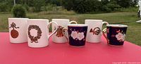 Photo showing six china mugs on red table with outdoor background, four white Christmas-themed mugs and two dark blue floral motif mugs.