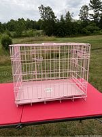 Pink pop up pet kennel placed on a red table outdoors, showing wire mesh construction with door latches and plastic tray base.