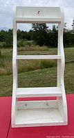 Photo of white wooden 3-tier shelf showing overall structure with curved side supports and flat shelves against outdoor background.