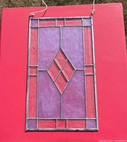Photo of rectangular stained glass panel showing geometric layout with purple and red textured glass and star-etched diamond center, metal framing with hanging loops.