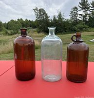 Photo showing three vintage glass jugs on red surface outdoors. Two amber glass jugs, one with a handle and one without, and one clear glass jug in the center.
