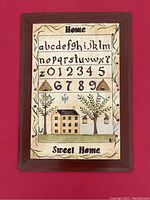 Framed folk art sampler with embroidered alphabet, numbers, and country house design on fabric background.