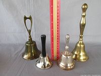 Four hand bells in a line with a red ruler behind them showing their relative heights. The bells vary in size and handle style.