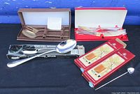 Overview of bronze utensils in box, large silver plated serving spoon, and two boxed sets of Birks drink stir spoons on black cloth