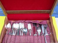 Photo of the cutlery stored in a classic wooden chest lined with red fabric, showing a mix of teaspoons, tablespoons, forks, knives, and serving utensils arranged in fitted fabric slots.