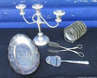 Full view of silver plate tableware including candelabra, tray, coasters, tongs, and cheese slicer against black cloth and blue wall