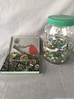 Overview of Meccano pieces in open Players cigar tin alongside a clear teddy money bank filled with marbles