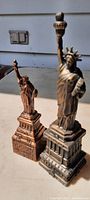 Two vintage metal Statue of Liberty figures, one bronze-colored approximately 8 inches tall, and one grayish approximately 9 inches tall, positioned side by side.