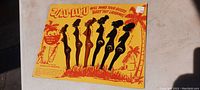 Front view of the original vintage cardstock display with six Zulu Lulu plastic drink stirrers attached. Yellow-orange cardstock with palm tree and tropical themed design and text.