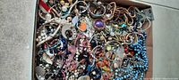 Wide view of flat box filled with mixed costume jewelry pieces showing variety of beads, bangles, and chains