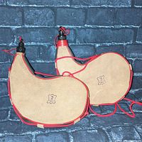 Two beige suede-like crescent-shaped bota bags with red trim and black plastic nozzles, each marked 2L Made in Spain.
