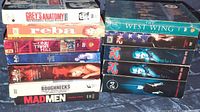 Photo of 11 TV series DVD box sets stacked in two piles, titles visible including Grey's Anatomy, Reba, One Tree Hill, 24, Roughnecks, Mad Men