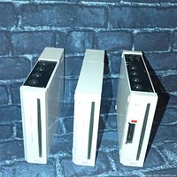 Front and side view of three vertical white Nintendo Wii consoles arranged side-by-side on a blue brick background. Each console shows disc slot and top of console.