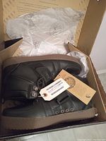 Top angled view of the left shoe in the open branded box showing black material, sole, and attached tags.