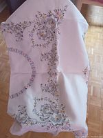 Photo showing hand embroidered floral patterns at the corner and edges of a cream or off-white linen square tablecloth.