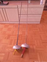 Pair of vintage epee fencing swords laid side by side on wooden floor with cabinets in background, showing full length and overall condition.