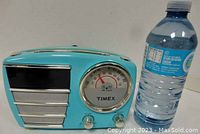 Front view of the turquoise Timex AM/FM clock radio with analog clock face and controls visible, next to a water bottle for size reference.