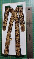 Leopard print suspenders laid flat on white cardboard with stainless steel ruler showing length and width, metal clips visible.