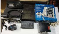 Box lot of various electronics showing a vintage Uniden CB radio, amplified phone in box, and multiple black electronics units on a shelf.