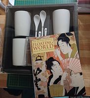 Overhead view of set showing four white ceramic tumblers, four long white mixing spoons, four brown sleeves, and the included book with illustrated cover.