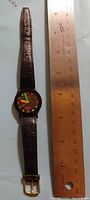 Front view of Jordache wristwatch on a ruler showing size. Watch has black round face with multicolor hands and dots as hour markers. Dark brown leather strap with texture and gold-tone buckle.