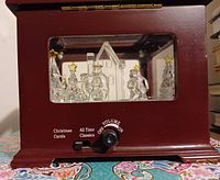 Close-up view of front panel showing volume knob and music selection switch labeled Christmas Carols and All Time Classics, inside are crystal figurines of Christmas trees and angels around a crystal church under a glass panel in mahogany wood casing.