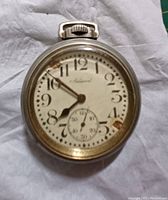 Front view of vintage Admiral pocket watch on a white cloth showing rust and missing stones on the dial, with visible hands and sub-second dial.
