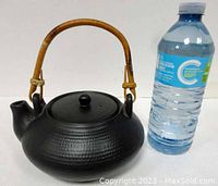 Front view of black ceramic teapot with bamboo handle alongside water bottle for size reference