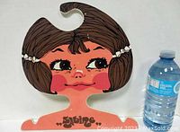 Front view of painted pop art hanger with stylized girl's face and hair, shown next to water bottle for scale.