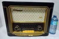 Front view of vintage Grundig Classic Model 960 radio, showing black Bakelite casing with gold trim, cream speaker grille, tuning dials, and yellow push buttons. Water bottle present for size reference.