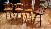 Four wooden swivel bar stools arranged side by side on a wooden floor, showing overall appearance and condition.