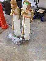 Photo shows two blow mold angels standing on a concrete floor, holding gold trumpets, and a silver-gray blow mold farm animal figure laying down in front of them.