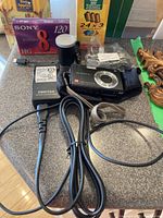 Photo showing Pentax Optio A30 camera, charger with power cord, battery, wrist strap, and multiple film packs along with calculator and pen.